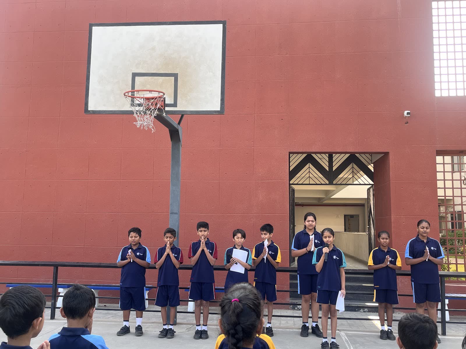 AARK students standing in a line with folded hands during morning assembly in the red-walled courtyard